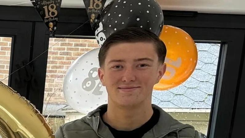 Henry Nowak in an olive green jacket smiles at the camera with 18th birthday balloons and banners behind him