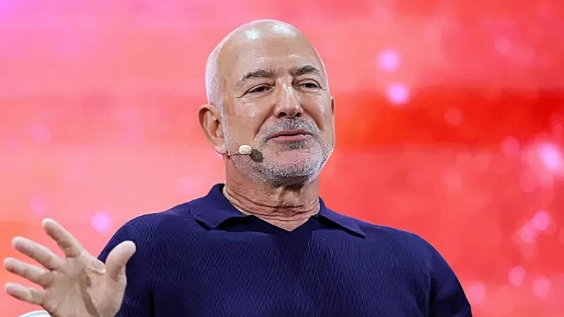 Reuters Jeff Bezos wearing a headset microphone and a navy blue polo shirt. He is on stage in front of a bright red background