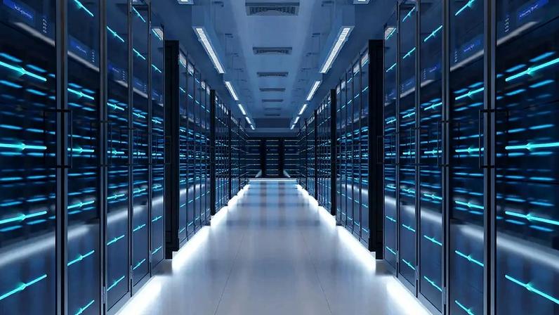 Corredor de um data center com fileiras de racks de servidores pretos, exibindo luzes azuis brilhantes que indicam atividade. O chão e o teto são claros, com iluminação linear, criando uma perspectiva futurista e tecnológica