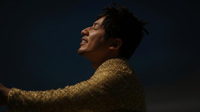 Jon Batiste in a gold sequined outfit sits on concrete looking up at a sunset sky