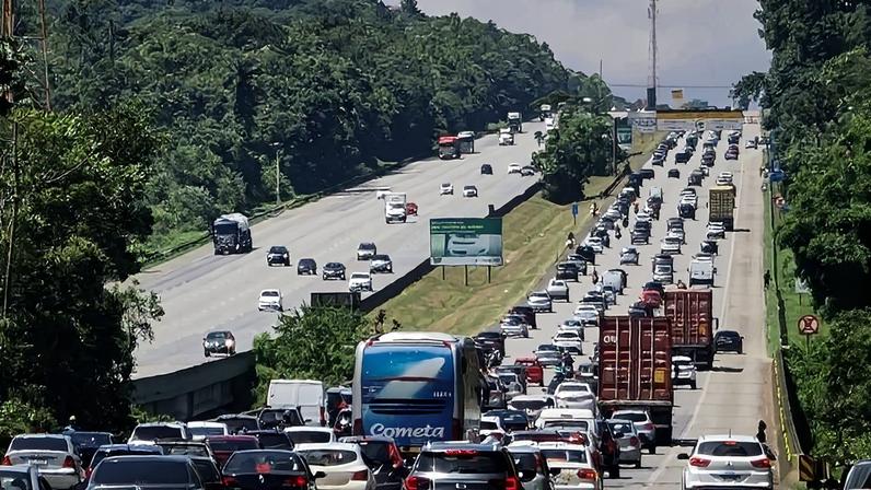 Imagem de uma rodovia com duas pistas largas. A pista da direita está completamente cheia de carros. Na outra, poucos veículos andam em sentido contrário. Nas laterais, muitas árvores cobrem os espaços.