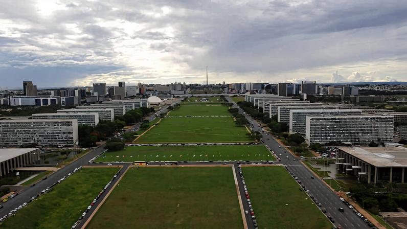 Vista panorâmica do Eixo Monumental em Brasília com grandes edifícios simétricos dos dois lados e uma ampla área verde central. O céu está nublado e a perspectiva mostra a extensão da avenida até o horizonte.