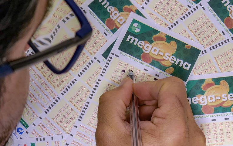 Pessoa com óculos segura caneta e marca números em cartão de aposta da Mega-Sena. Vários cartões semelhantes estão espalhados sobre a mesa.