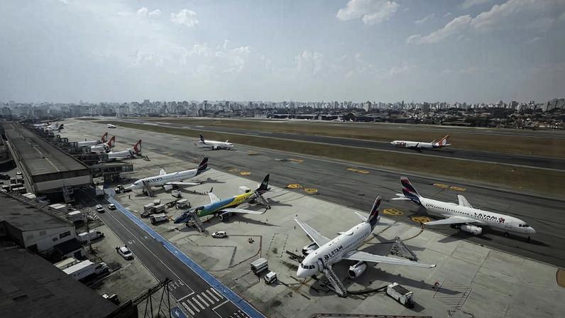Quatro aviões estão estacionados em posições próximas a um terminal de aeroporto. O terminal possui pontes de embarque conectadas às aeronaves. Ao fundo, há pista de pouso e decolagem com área urbana visível no horizonte sob céu parcialmente nublado.