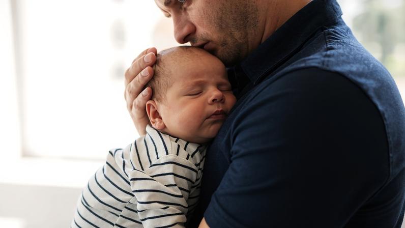 Um homem está segurando um bebê adormecido em seus braços. O bebê está vestido com uma roupa listrada em branco e azul, enquanto o homem usa uma camisa escura. O ambiente é iluminado, sugerindo um espaço acolhedor e tranquilo.