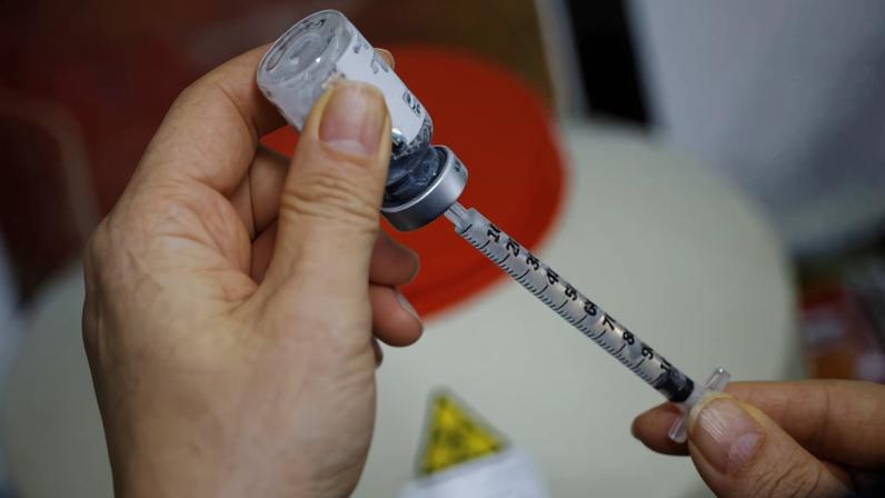 Uma mão segurando uma seringa com um frasco de medicamento. A seringa está parcialmente preenchida e o frasco está sendo retirado. Ao fundo, há um recipiente com tampa vermelha e um símbolo de alerta.