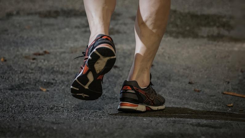 A imagem mostra as pernas de uma pessoa em movimento, com foco nos pés calçados com tênis de corrida. O chão é de asfalto e há algumas folhas secas ao redor. O tênis é predominantemente escuro com detalhes em laranja e branco.