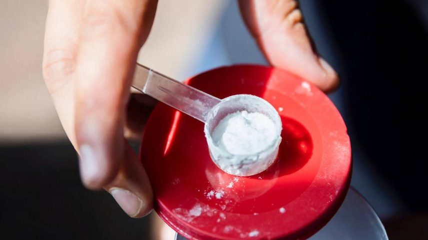 Uma mão segurando uma colher medidora transparente com um pó branco dentro, posicionada sobre uma tampa vermelha. O fundo é desfocado, mas sugere um ambiente ao ar livre.