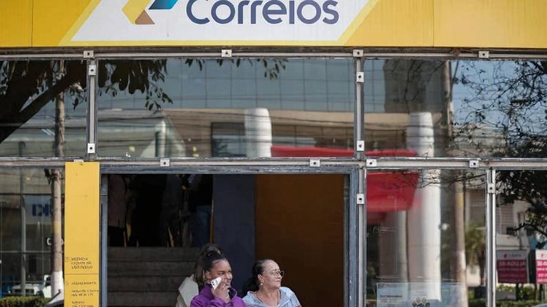 A imagem mostra a entrada de uma agência dos Correios, com um letreiro amarelo e azul. Duas mulheres estão saindo da agência. A mulher à esquerda está usando um moletom roxo e segurando um celular, enquanto a mulher à direita está vestindo uma blusa clara e segurando um papel. Ao fundo, é possível ver escadas e outras pessoas dentro da agência.