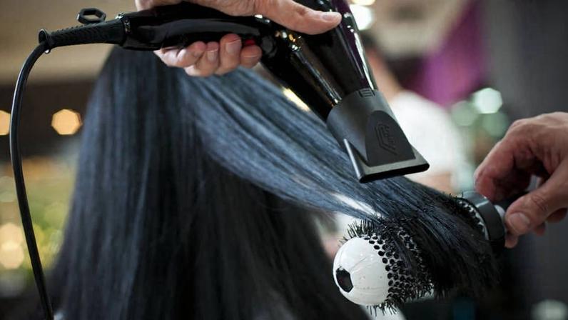 A imagem mostra uma pessoa secando o cabelo com um secador. O cabelo é longo e liso, e está sendo modelado com uma escova redonda. A mão que segura o secador é visível, assim como a escova que está em contato com o cabelo. O fundo é desfocado, com luzes suaves que sugerem um ambiente de salão de beleza.