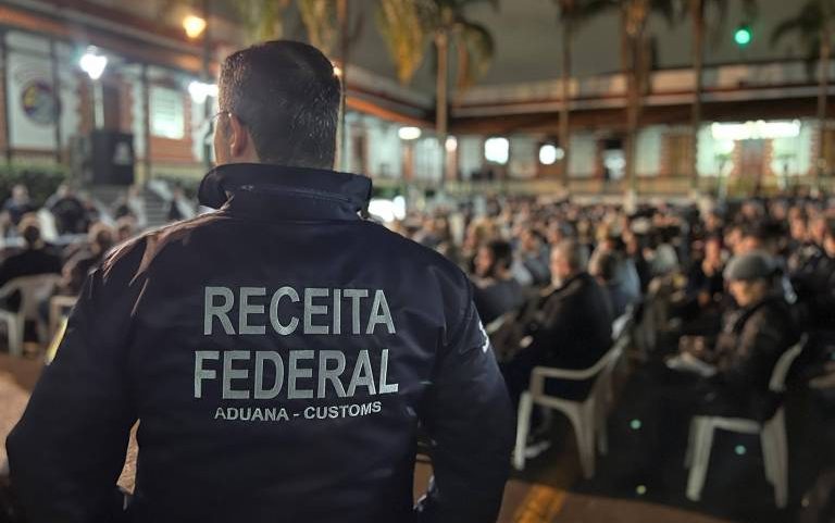 A imagem mostra um agente da Receita Federal, de costas, usando um uniforme escuro com a inscrição 'RECEITA FEDERAL' e 'ADUANA - CUSTOMS'. Ao fundo, há uma grande quantidade de pessoas sentadas em cadeiras brancas, em um ambiente iluminado à noite, com palmeiras visíveis ao redor.