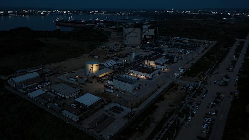 Vista aérea de complexo industrial com tanques de armazenamento e edifícios, cercado por vegetação. Ao fundo, navios atracados em porto marítimo sob céu claro.