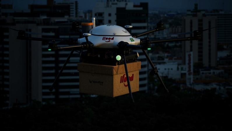 Drone multirotor com oito hélices transporta caixa de entrega com logotipo da empresa, voando acima de área verde com prédios residenciais e comerciais ao fundo.