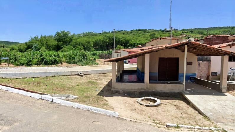 Casa térrea com varanda coberta em área rural, com duas pessoas sentadas na varanda. Vegetação verde e colinas ao fundo sob céu azul com nuvens.