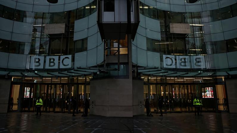 Fachada curva do edifício da BBC com logo iluminado acima da entrada principal. Portas de vidro refletem o logo e há seguranças com coletes amarelos na entrada.