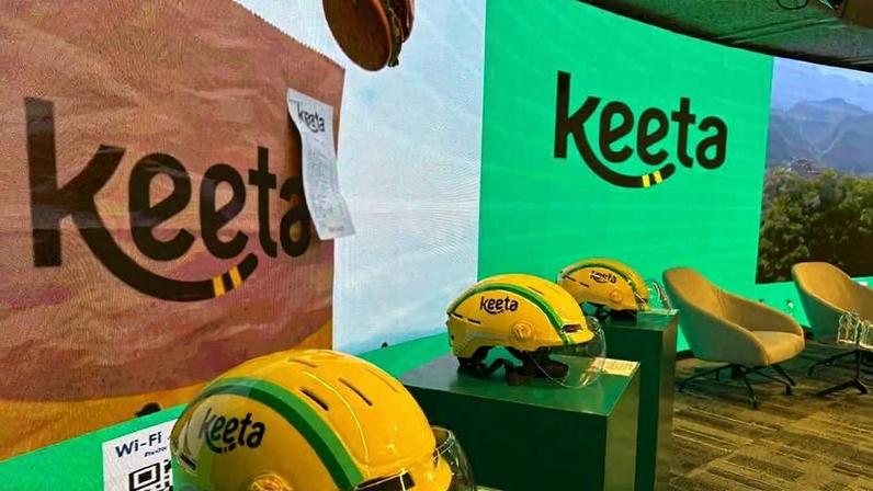 Três capacetes amarelos com o logo Keeta expostos sobre pedestais verdes em ambiente interno. Ao fundo, telas exibem o logo Keeta em verde e marrom, além de uma imagem de paisagem montanhosa.