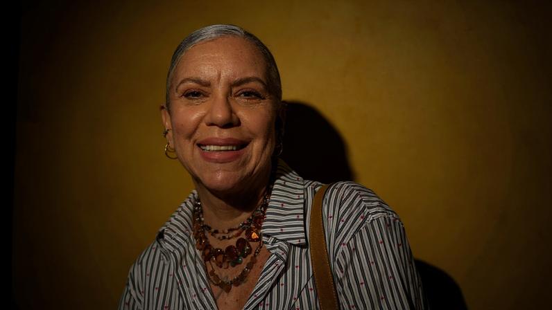 Mulher de meia-idade com cabelo grisalho preso, usando camisa listrada branca com detalhes vermelhos e colares coloridos. Ela sorri para a câmera contra fundo amarelo liso.