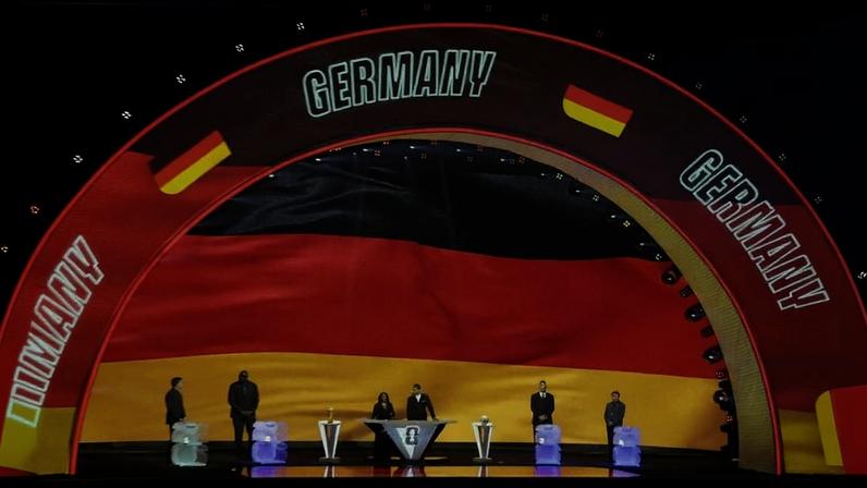 Seis pessoas em pé em palco iluminado com fundo da bandeira da Alemanha. Arco acima exibe a palavra "GERMANY" e bandeiras alemãs em destaque.