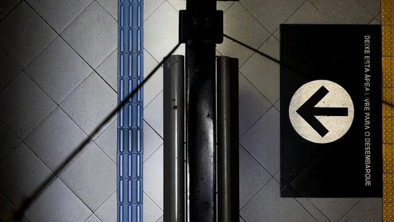 Vista aérea mostra duas pessoas na plataforma de metrô ao lado de um banco preto. Piso cinza com faixa tátil azul e placa preta com seta branca indicando direção para desembarque. Texto na placa orienta manter a esquerda para desembarque.