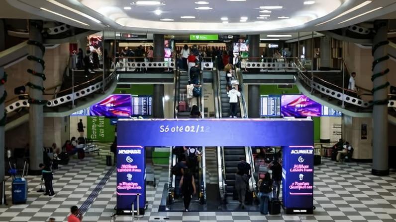 Interior do aeroporto de Congonhas, com piso quadriculado e escadas rolantes ao centro. Pessoas caminham e esperam, algumas com malas. Painéis digitais exibem informações e anúncios em roxo e branco.