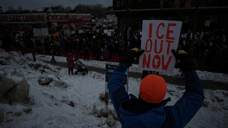 Pessoa com jaqueta azul e gorro laranja segura cartaz escrito 'ICE OUT NOW' em letras laranja durante protesto em área coberta de neve em Chicago. Multidão reunida em frente a loja Chicago Lake Liquors, com várias pessoas e carrinhos de bebê visíveis.