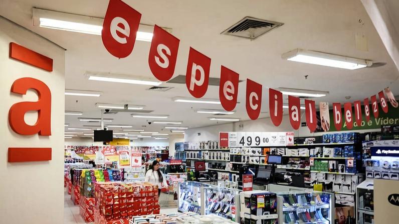 Interior de loja com decoração vermelha pendurada formando a frase 'especial beleza'. Prateleiras exibem eletrônicos, acessórios e produtos variados. Cliente caminha entre os corredores iluminados.