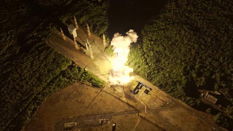 Foguete é lançado de plataforma iluminada à noite, com chama intensa e fumaça visível. Área ao redor é coberta por vegetação densa e algumas estruturas próximas.
