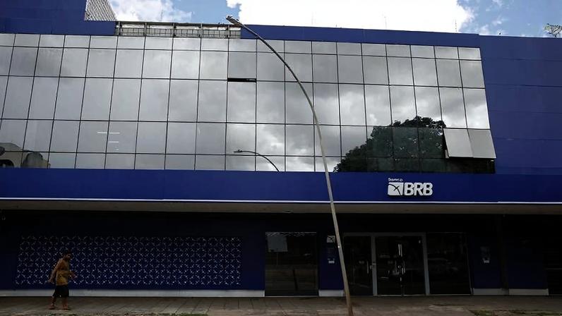 Fachada azul do banco BRB com janelas espelhadas refletindo o céu e árvores. Homem caminha na calçada em frente ao prédio.