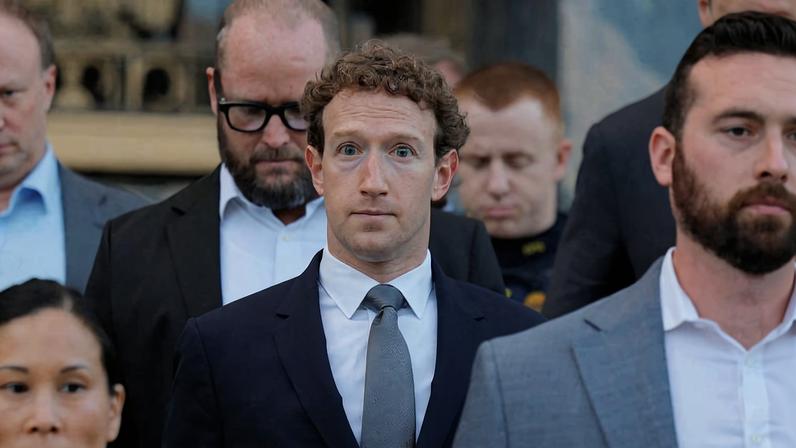 Mark Zuckerberg veste terno escuro e gravata cinza, olhando diretamente para frente. Ele está cercado por outras pessoas em trajes formais, algumas olhando para baixo, em ambiente interno.