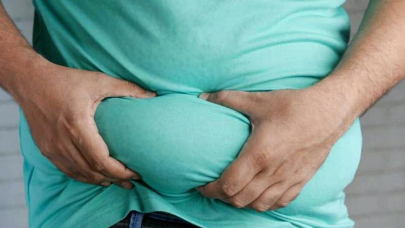 Pessoa vestindo camiseta azul clara segura a barriga com as duas mãos, apertando a região abdominal. A imagem mostra o tronco da pessoa, com parte da calça jeans e cinto visíveis.