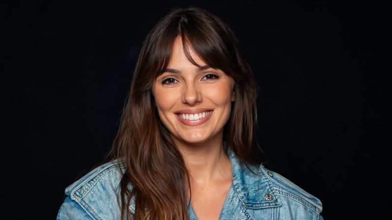 Mulher jovem com cabelo longo e liso veste jaqueta jeans azul clara e está sentada em cadeira de madeira, sorrindo para a câmera. Ao fundo, outras pessoas estão sentadas em arquibancada colorida.