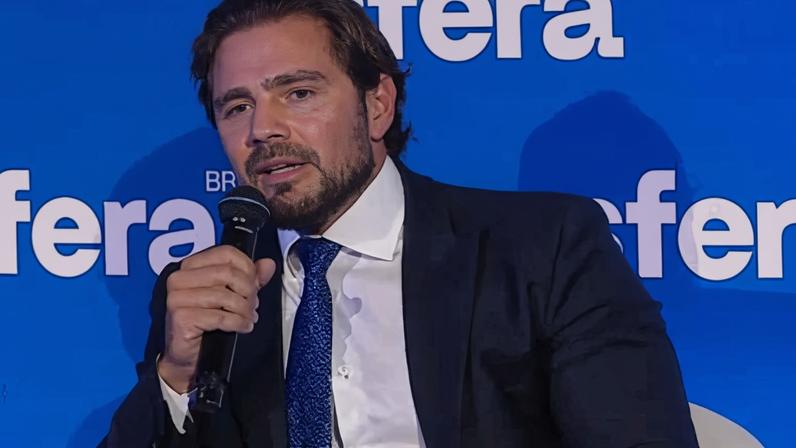 Homem de terno escuro e gravata azul segura microfone enquanto fala sentado em cadeira branca. Fundo azul exibe texto branco 'fera BR'.