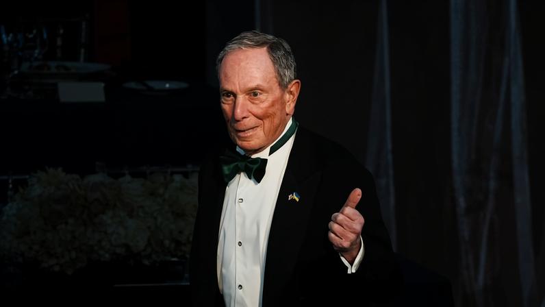 Homem idoso veste smoking preto com gravata borboleta verde e camisa branca, fazendo sinal de positivo com a mão direita. Ao fundo, mesa decorada com arranjo de flores brancas e rosas, e utensílios de jantar.