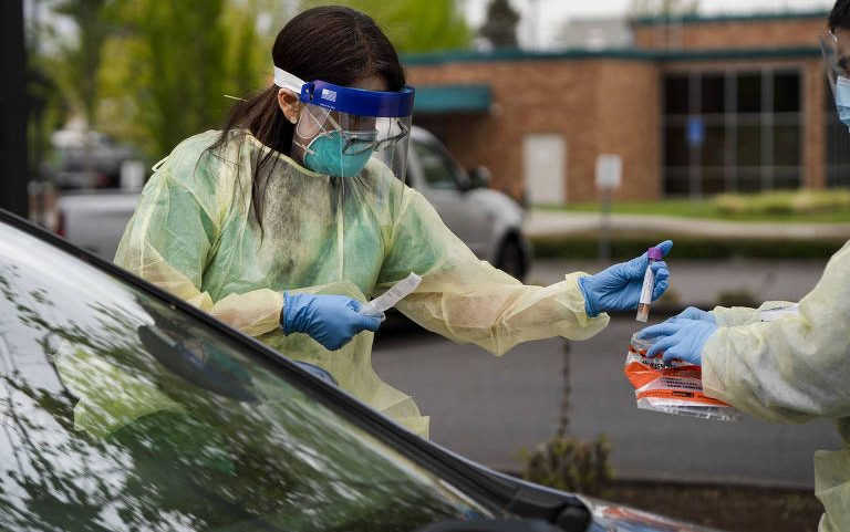 Profissional de saúde com máscara, viseira e avental coleta amostra de motorista em teste de Covid-19 ao ar livre. Ambiente externo com árvores e prédio ao fundo.