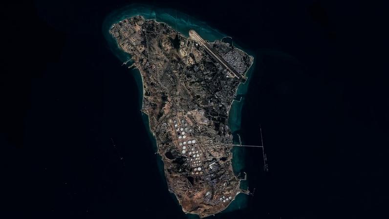 Imagem aérea mostra ilha com instalações industriais, tanques de armazenamento e pista de pouso, cercada por mar azul.