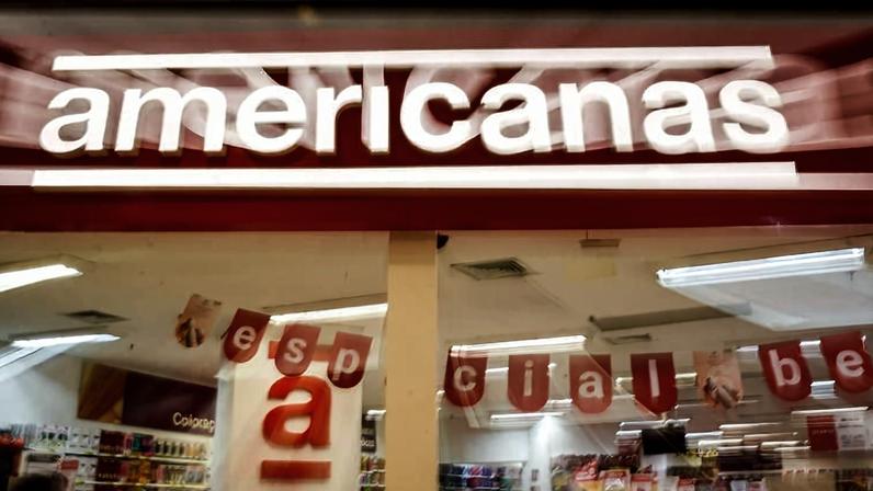Fachada da loja Americanas com letreiro iluminado em vermelho e branco. No interior, decoração com letras vermelhas penduradas formando palavras, prateleiras com produtos e clientes circulando.
