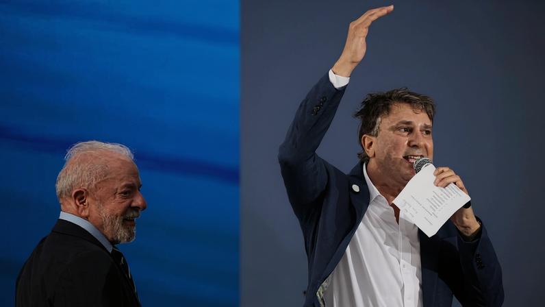Homem de terno azul e camisa branca fala ao microfone com a mão direita levantada, segurando papéis na esquerda. Outro homem de cabelos brancos e barba observa sorrindo ao lado, ambos em frente a fundo azul e branco.