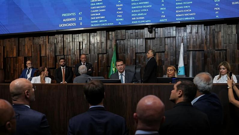 Dez parlamentares sentados e em pé em plenário com painel eletrônico ao fundo mostrando presença e nomes. Pessoas observam a sessão em primeiro plano.