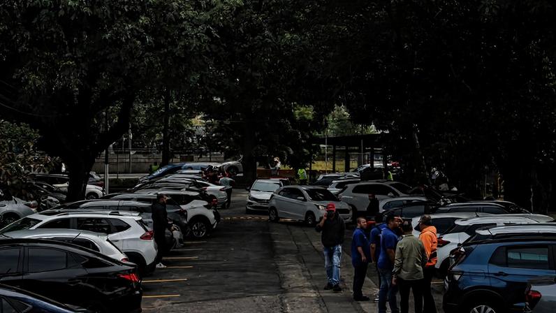 Estacionamento ao ar livre com vários carros estacionados em filas sob árvores. Grupo de seis pessoas conversa próximo a um carro azul à direita. Ambiente urbano com vegetação densa ao redor.