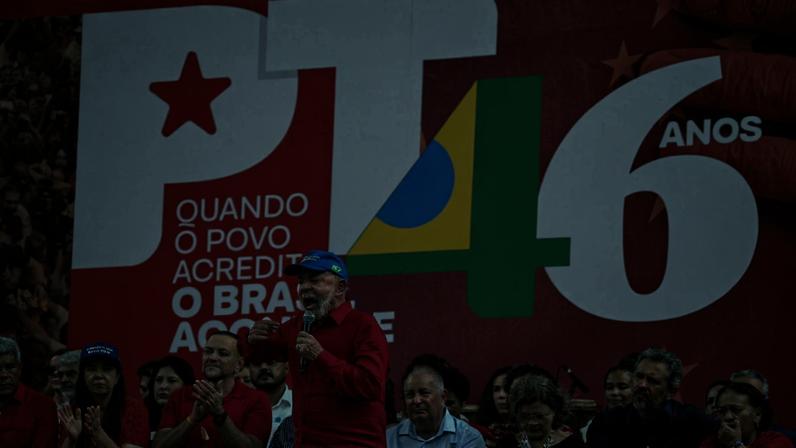 Líder do PT fala ao microfone em comício com fundo vermelho e logo do PT com número 46 em verde e amarelo. Pessoas ao redor vestem vermelho e aplaudem.