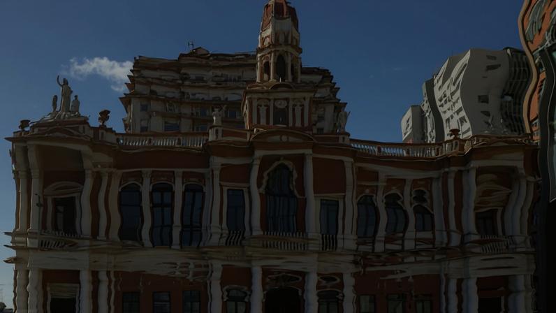 Reflexo ondulado de um prédio antigo com arquitetura clássica, incluindo colunas e janelas altas, visto em superfície de água que distorce a imagem. Céu azul com poucas nuvens ao fundo.