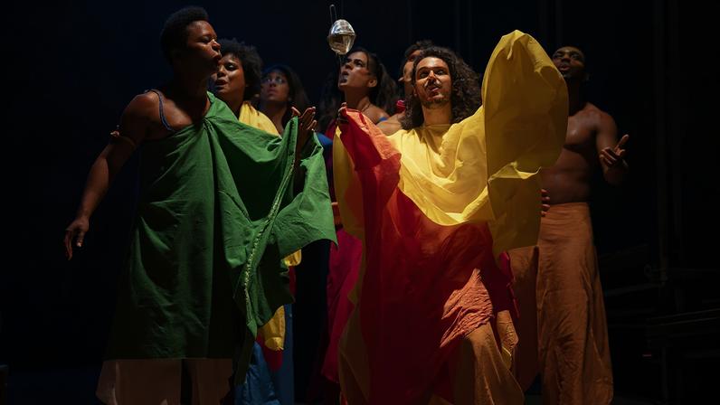 Dez pessoas em palco escuro participam de performance teatral, vestindo tecidos coloridos em tons vibrantes como verde, amarelo e vermelho, iluminadas por luzes vermelhas ao fundo.