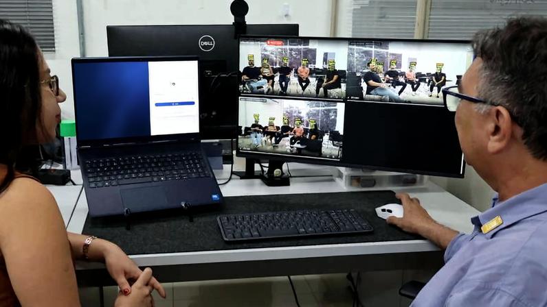 Homem e mulher sentados lado a lado em mesa com laptop e dois monitores. Laptop exibe imagem colorida de peças de quebra-cabeça, enquanto monitores mostram várias janelas de videoconferência com grupos de pessoas em ambiente interno.