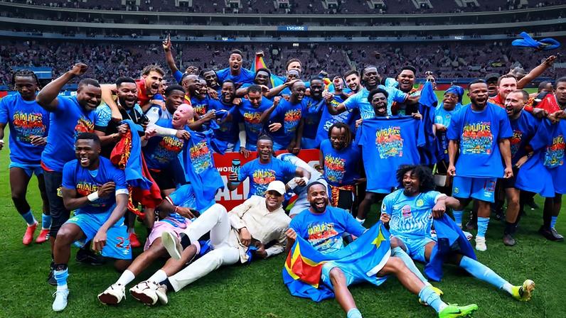 Grupo de jogadores de futebol e equipe técnica posam juntos no gramado de estádio lotado, usando camisetas comemorativas azuis. Alguns seguram bandeiras e exibem sorrisos, com arquibancadas vermelhas e público ao fundo.