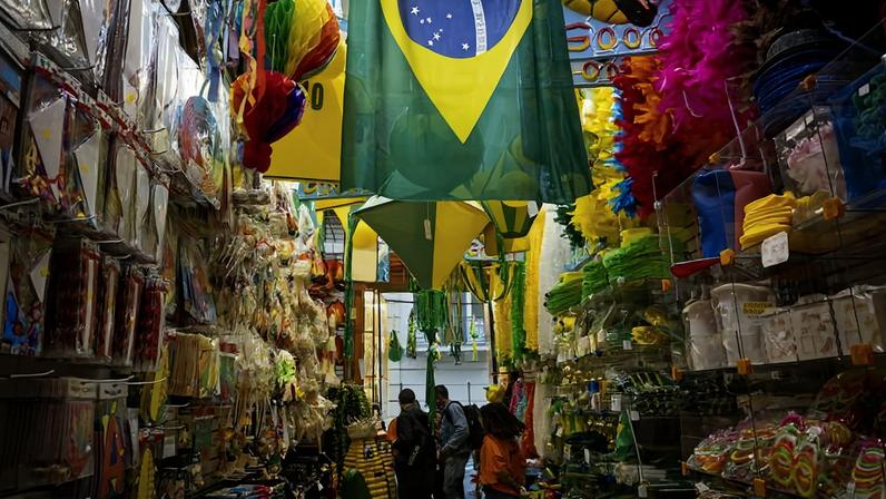 Interior de loja com decoração e produtos nas cores verde e amarelo, incluindo bandeiras do Brasil penduradas no teto. Prateleiras cheias de artigos esportivos, fantasias e acessórios relacionados ao futebol. Três pessoas observam os produtos no corredor central.