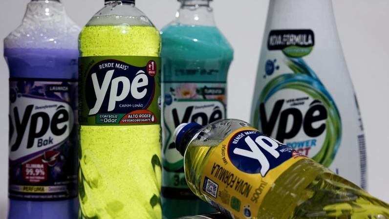 Cinco garrafas plásticas de produtos de limpeza da marca Ypê em diferentes cores e rótulos. Em destaque, uma garrafa amarela com rótulo preto e azul, ao centro, e outras garrafas nas cores roxa, verde, branca e amarela ao redor, algumas em pé e uma deitada.