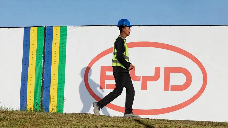 Homem usando capacete azul e colete refletivo caminha sobre grama em frente a muro branco com logo vermelho da BYD e faixas verticais nas cores azul, verde e amarelo.