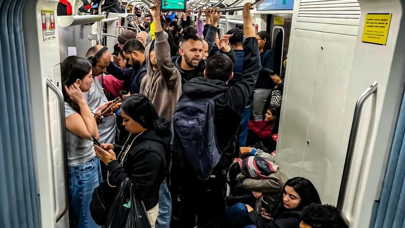 Vagão de trem cheio com passageiros em pé segurando barras e outros sentados no chão e bancos laterais. Alguns usam mochilas e fones de ouvido, e vários estão concentrados em seus celulares. Ambiente interno com iluminação artificial e paredes claras.