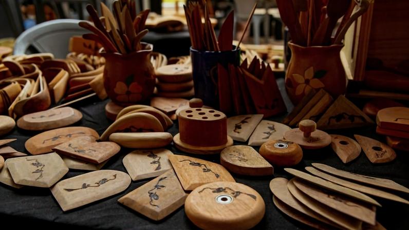 Mesa com diversos objetos artesanais de madeira, incluindo porta-copos, suportes, e utensílios decorados com desenhos simples. Alguns itens estão organizados em vasos pintados com flores, e outros dispostos diretamente sobre a mesa coberta por tecido preto.