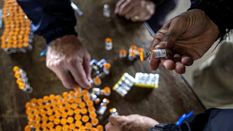 Quatro mãos manipulam frascos pequenos com tampas laranjas sobre mesa com vários frascos semelhantes organizados em grupos. Ambiente interno com foco nos frascos e mãos.